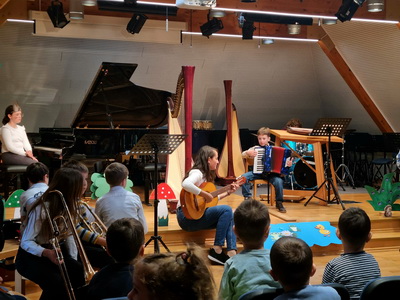 Glasbena pravljica v glasbeni šoli – učiteljica igra na kitaro, deček na harmoniko, v ospredju otroci